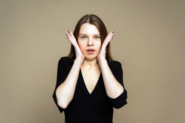 Şok olmuş bir kız omuz silkiyor. Kahverengi arka planda bir kız var. Yüzünde elleri olan bir kız.