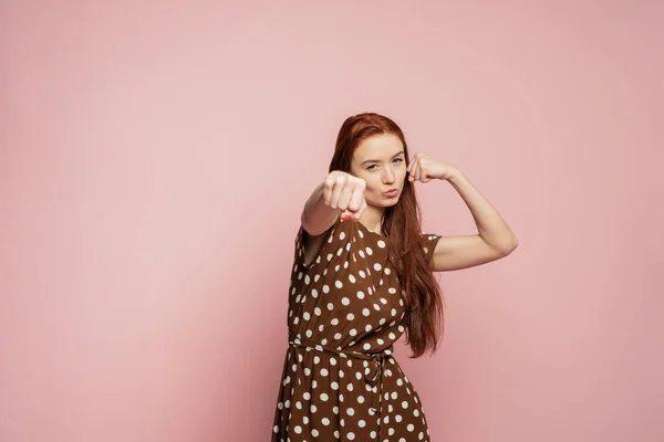 A redhead girl is on the pink background doing sport. A woman is boxing . A strong girl is ready to hit.
