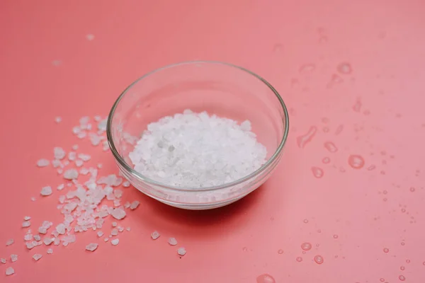 sea salt in a glass bowl on a pink pastel background next to sea water.