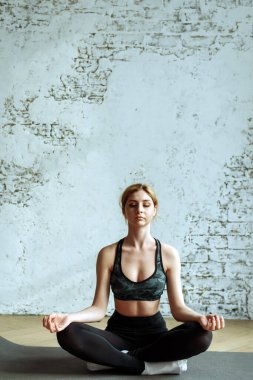 Güzel bir genç kız, evde, nilüfer çiçeğinin içinde meditasyon yapmakla meşgul. Tatlı sarışın beyaz tuğlalı bir duvarın arka planında fitness yapıyor..
