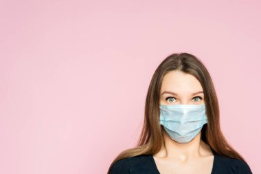 Pembe arka planda koruyucu maskeli korkmuş genç bir kadın. Koronavirüs salgınının korkusu ve dehşeti.