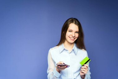 Gülümseyen bir kız internetten mavi arka planda sipariş veriyor. Cep telefonu ve kredi kartı olan bir kız..