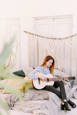 Yetişkin bir kadın boho tarzı bir odada yatağın üzerinde gitar çalar. Güzel ve rahat bir atmosfer..