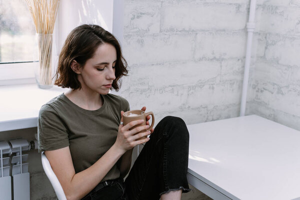 Beautiful young woman basking on a cup of coffee or tea in her bright kitchen.