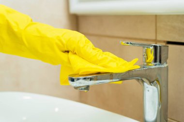 Birisi sarı eldivenlerle lavaboyu yıkıyor. Birisi banyoyu deterjanla temizliyor..