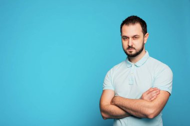 Mavi arka planda şüpheli bir adam var. Kollarını kavuşturmuş sakallı bir adam. Sakallı bir adam şüpheli görünür..