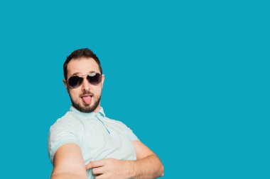 Genç, sakallı bir adam mavi arka planda kameraya bakıyor. Adam bir selfie çekip komik suratlar yapıyor..