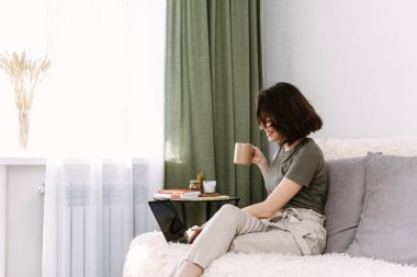 Yatak odasındaki dizüstü bilgisayarla bir fincan sıcak kahve ya da çay içen genç bir kadın. Kadın sosyal uygulamaları kontrol ediyor, online alışveriş yapıyor ve çalışıyor. İletişim ve teknoloji kavramı.