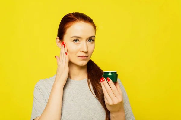 Una chica pelirroja está aplicando crema en la cara. Una chica unta crema facial sobre el fondo amarillo — Foto de Stock