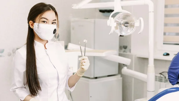 Vrolijke vrouwelijke tandarts glimlachend op haar kantoor. Mooie vrouw student in een beschermend masker in de tandheelkunde staat in de tandheelkundige kantoor en houdt professionele gereedschappen — Stockfoto