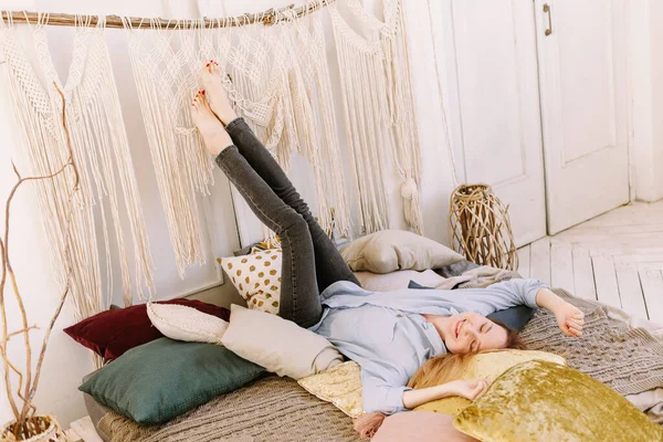 Genç beyaz bir anne yatakta uzanıyor ve ayaklarını beyaz duvara doğru kaldırıyor. Yetişkin, sevimli bir kadın evde boho tarzında yatıyor.