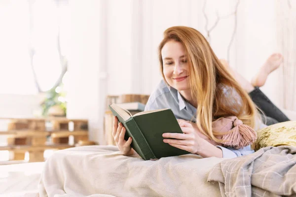 Милая женщина, читающая книгу, лежащая в постели. Молодая взрослая девушка учится дома одна — стоковое фото