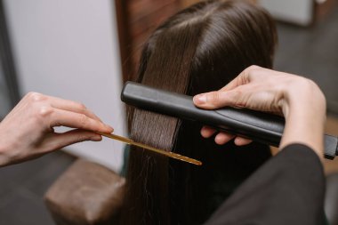 Kuaför bir saç tarağı ve ütüleme ile bir güzellik salonunda bir esmerin saçını çeker ve düzeltir. Keratin geri kazanma saç ve protein yığını profesyonel ultrasonik demir alet ile.