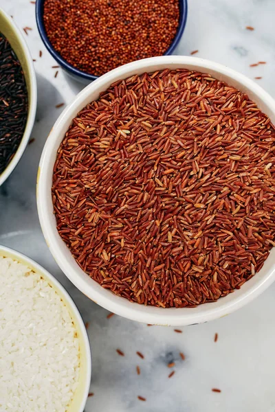 Ceramic bowls filled with a variety of grain and rice including red quinoa, red rice, black rice, and white rice on a marble top.