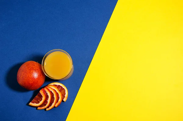 A whole red orange lies on a blue / yellow background. Slices of orange lie nearby and there is a glass of orange juice. Soft focus, top view. Trend blue background