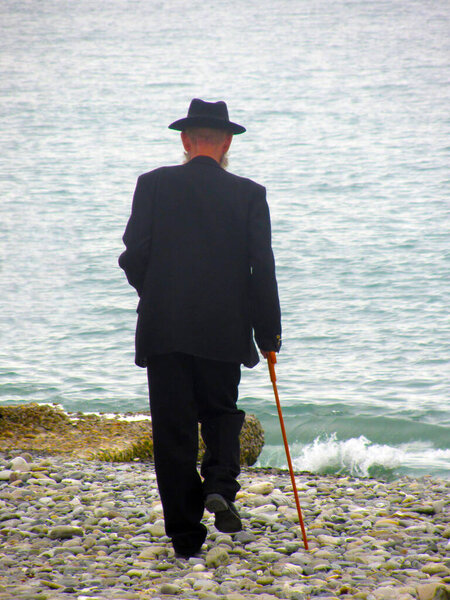 An elderly man in a black suit with a cane walking along a pebble beach to the sea. The concept of loneliness, retirement, relationships with relatives, disease, illness, sickness, ailment, death.