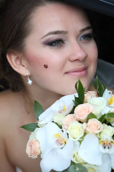 Seorang pengantin dengan karangan bunga di tangannya dan seekor ladybird di pipinya di dalam mobil di kursi belakang . — Stok Foto