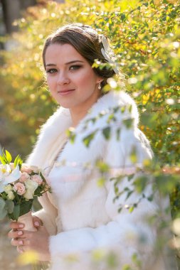 Beyaz kürklü gelin çalıların arkasında elinde bir düğün buketi tutuyor..