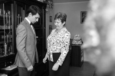 Anne, oğluyla damadın gelinlik giymesine yardım ediyor. Damat ve anne düğün için hazırlanıyorlar..