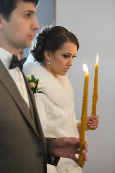 Bruid en bruidegom staan op huwelijksceremonie. Gelukkige stijlvolle bruiloft paar met kaarsen met licht onder gouden kronen tijdens het heilig huwelijk in de kerk. — Stockfoto