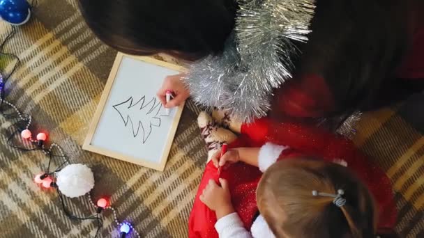 Anya a lányával a táblán karácsonyi mutatványokat rajzol. Anya és lánya karácsonyfát rajzolnak. Karácsonyi téma: anya és lánya festeni Karácsony egy otthonos ünnepi környezetben. — Stock videók