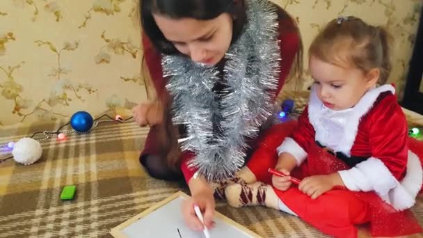 Mãe com filha no quadro desenhar atributos de Natal. Mãe e filha estão desenhando uma árvore de Natal. Tema de Natal: mãe e filha pintam o Natal em um ambiente festivo caseiro . — Vídeo de Stock