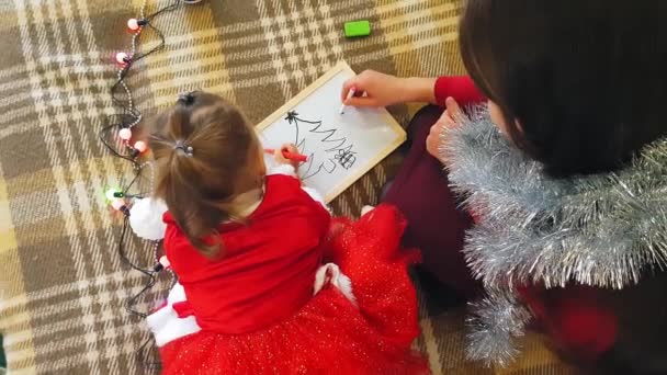 Η μαμά με την κόρη στο συμβούλιο ζωγραφίζουν τα Χριστουγεννιάτικα χαρακτηριστικά. Η μαμά και η κόρη ζωγραφίζουν ένα χριστουγεννιάτικο δέντρο. Χριστουγεννιάτικο θέμα: μαμά και κόρη ζωγραφίζουν τα Χριστούγεννα σε ένα σπιτικό εορταστικό σκηνικό. — Αρχείο Βίντεο
