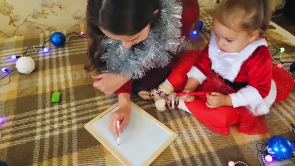 ボード上の娘とお母さんはクリスマスの属性を描きます。母と娘はクリスマスツリーを描いている。クリスマスのテーマ:お母さんと娘は家庭的なお祝いの設定でクリスマスをペイント. — ストック動画