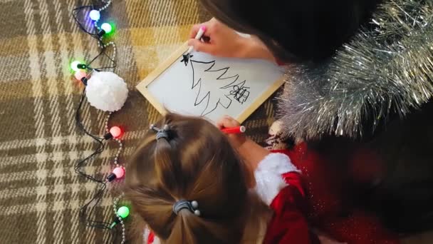 Anya a lányával a táblán karácsonyi mutatványokat rajzol. Anya és lánya karácsonyfát rajzolnak. Karácsonyi téma: anya és lánya festeni Karácsony egy otthonos ünnepi környezetben. — Stock videók