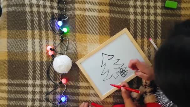 Mamá con hija en la pizarra dibuja atributos navideños. Mamá y su hija están dibujando un árbol de Navidad. Tema de Navidad: mamá e hija pintan Navidad en un ambiente festivo hogareño . — Vídeos de Stock