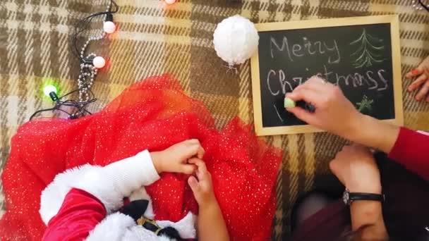 Mamá con hija en la pizarra dibuja atributos navideños. Mamá y su hija están dibujando un árbol de Navidad. Tema de Navidad: mamá e hija pintan Navidad en un ambiente festivo hogareño . — Vídeos de Stock