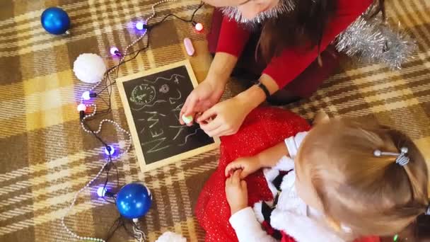 Mam en dochter tekenen nieuwjaarsattributen op het schoolbord. Mam en dochter tekenen een kerstboom. Nieuwjaarsthema: moeder en dochter schilderen het nieuwe jaar in een feestelijke sfeer. — Stockvideo