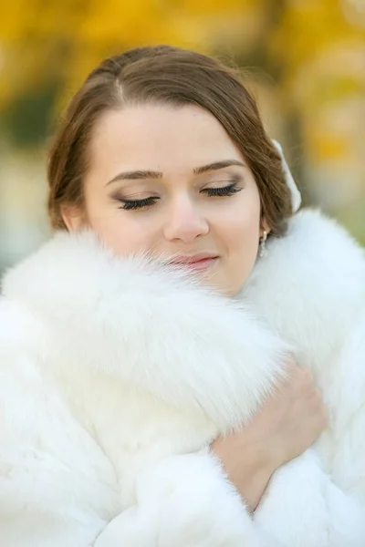 Bride with a bouquet of flowers in an autumn park in a white fur coat — Stock Photo, Image