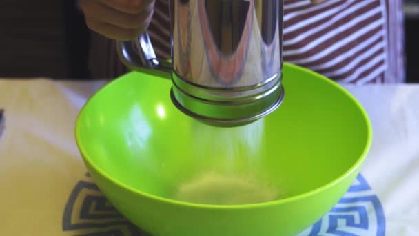 Close-up câmera lenta de uma mão feminina peneirando farinha com uma caneca peneira em uma tigela verde na cozinha da casa. Panquecas de cozinha — Vídeo de Stock