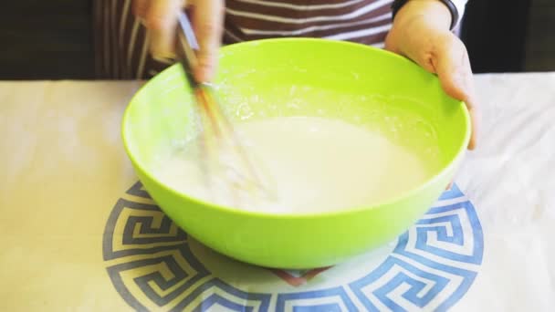 Крупный план женской руки смешивает тесто с кухонным венчиком в зеленой миске на домашней кухне. Блинчики для готовки — стоковое видео