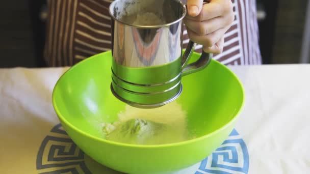 Close-up de uma mão feminina peneirando farinha com uma caneca peneira em uma tigela verde na cozinha da casa. Panquecas de cozinha — Vídeo de Stock