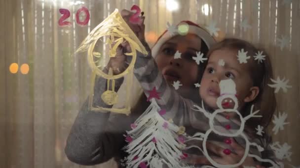 Mãe e filha desenhar um boneco de neve, uma árvore de Natal e um relógio com tintas em um painel de janela. Desenho com acrílicos em vidro . — Vídeo de Stock