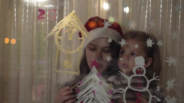 Η μαμά και η κόρη ζωγραφίζουν έναν χιονάνθρωπο, ένα χριστουγεννιάτικο δέντρο και ένα ρολόι με μπογιές σε ένα τζάμι παραθύρου. Σχέδιο με ακρυλικά σε γυαλί. — Αρχείο Βίντεο