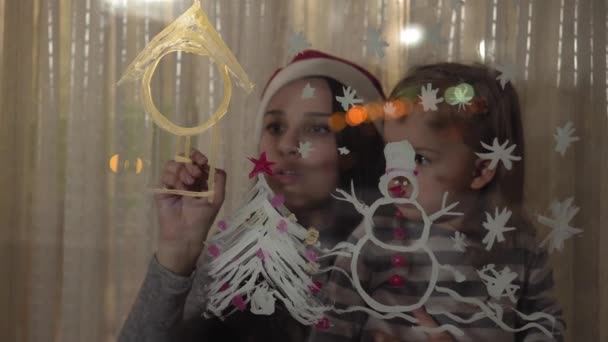 Mamma och dotter ritar en snögubbe, en julgran och en klocka med färger på en fönsterruta. Ritning med akryl på glas. — Stockvideo