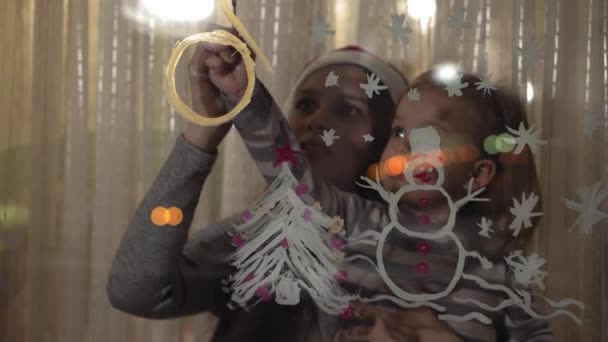 Mamma och dotter ritar en snögubbe, en julgran och en klocka med färger på en fönsterruta. Ritning med akryl på glas. — Stockvideo