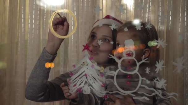 Mãe e filha desenhar um boneco de neve, uma árvore de Natal e um relógio com tintas em um painel de janela. Desenho com acrílicos em vidro . — Vídeo de Stock