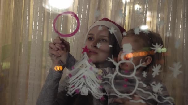 Mam en dochter tekenen een sneeuwpop, een kerstboom en een klok met verf op een ruit. Tekening met acryl op glas. — Stockvideo