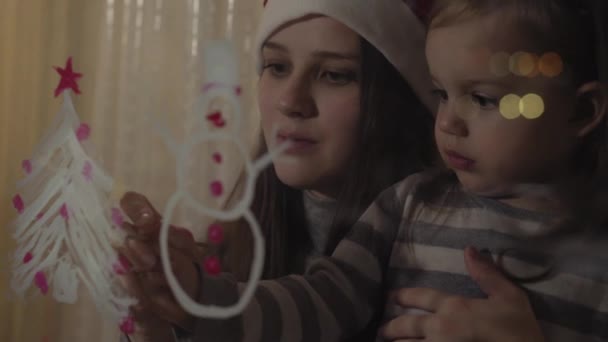 Mamá y su hija dibujan un muñeco de nieve y un árbol de Navidad con pinturas en el cristal de una ventana. Dibujo con acrílicos sobre vidrio . — Vídeos de Stock