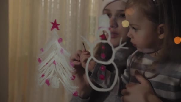 Mamá y su hija dibujan un muñeco de nieve y un árbol de Navidad con pinturas en el cristal de una ventana. Dibujo con acrílicos sobre vidrio . — Vídeos de Stock