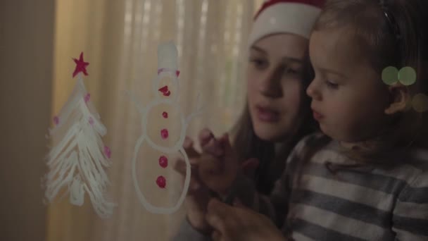 Η μαμά και η κόρη ζωγραφίζουν έναν χιονάνθρωπο και ένα χριστουγεννιάτικο δέντρο με μπογιές σε ένα τζάμι παραθύρου. Σχέδιο με ακρυλικά σε γυαλί. — Αρχείο Βίντεο