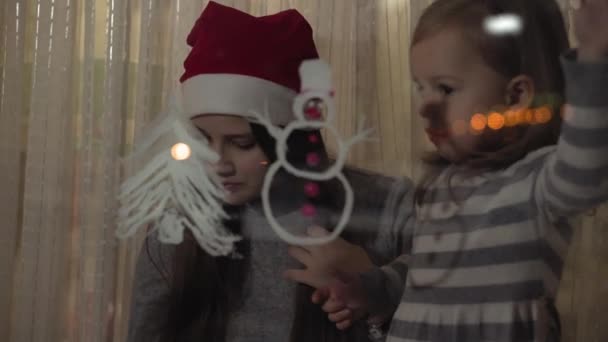 Mamá y su hija dibujan un muñeco de nieve y un árbol de Navidad con pinturas en el cristal de una ventana. Dibujo con acrílicos sobre vidrio . — Vídeos de Stock