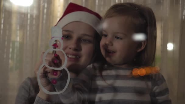 Maman et fille dessinent un bonhomme de neige avec de la peinture sur une vitre. Dessin avec acrylique sur verre . — Video