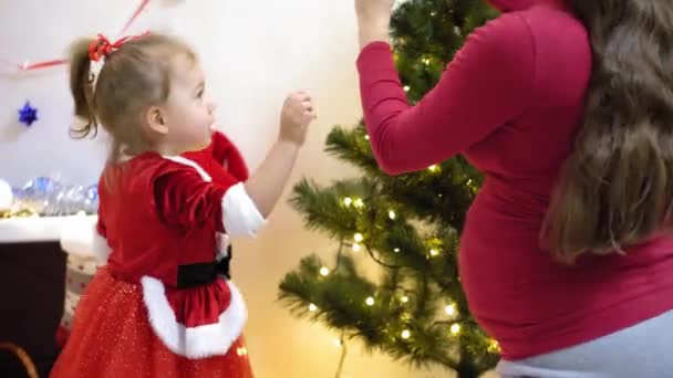 Baby och pregnan mamma hänga röd boll leksak på julgran. Glad barndom. barn och mor dekorera trädet med julbollar. litet barn och en förälder leker vid julgranen. — Stockvideo