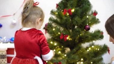 Bebek ve hamile bir anne Noel ağacına kırmızı top oyuncağı asıyor. Mutlu çocukluk kavramı. Çocuk ve anne Noel toplarıyla ağacı süslerler. Küçük çocuk ve bir ebeveyn Noel ağacının yanında oynuyorlar..