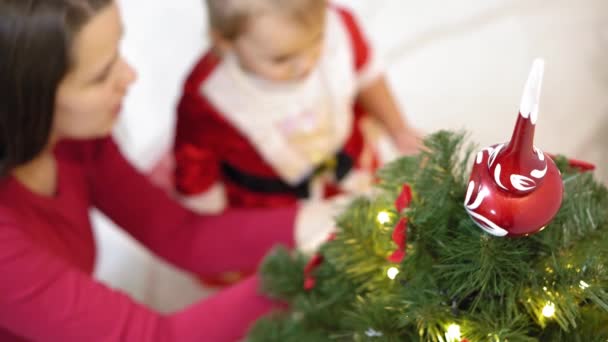 엄마와 아기가 크리스마스 트리에 빨간 공 장난감을 걸었어요. 행복 한 어린 시절의 컨셉. 아이와 엄마가 크리스마스 때 공으로 나무를 장식하는 거야. 어린 아이와 부모님이 크리스마스 트리에서 연주를 하고 있습니다. — 비디오